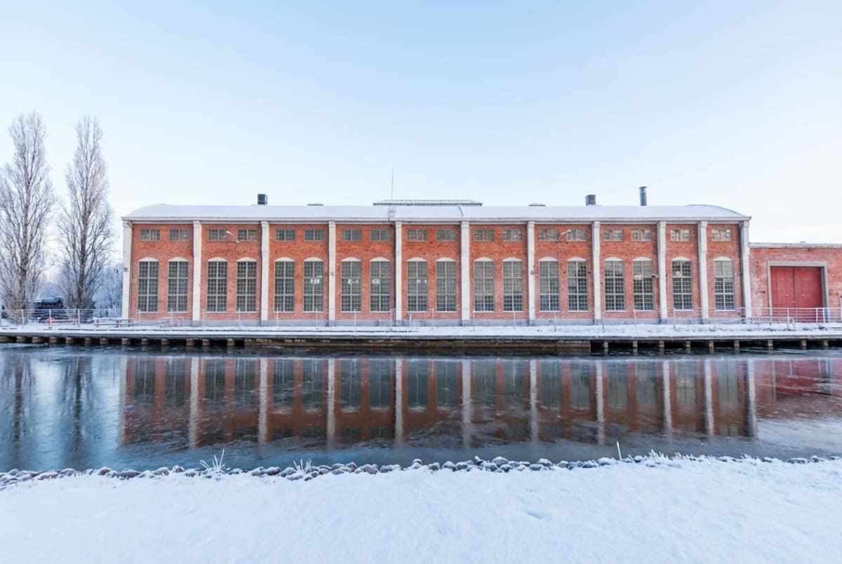 En fasadbild av en stor tegelbyggnad med många fönster längs en vattenkant en vinterdag. Byggnaden har en orange-röd tegelfasad och vita vertikala band som delar in fönsterraderna. Taket är platt och täckt med snö. Framför byggnaden ligger en vattenyta som reflekterar byggnaden, och vattnet verkar delvis fruset. Nära vattenkanten i förgrunden syns snöig mark med några stenar och vissna grässtrån. Till vänster om byggnaden står flera höga, kala träd täckta med snö. Himmel är ljusblå och klar. På byggnadens högra sida finns en röd industriport.