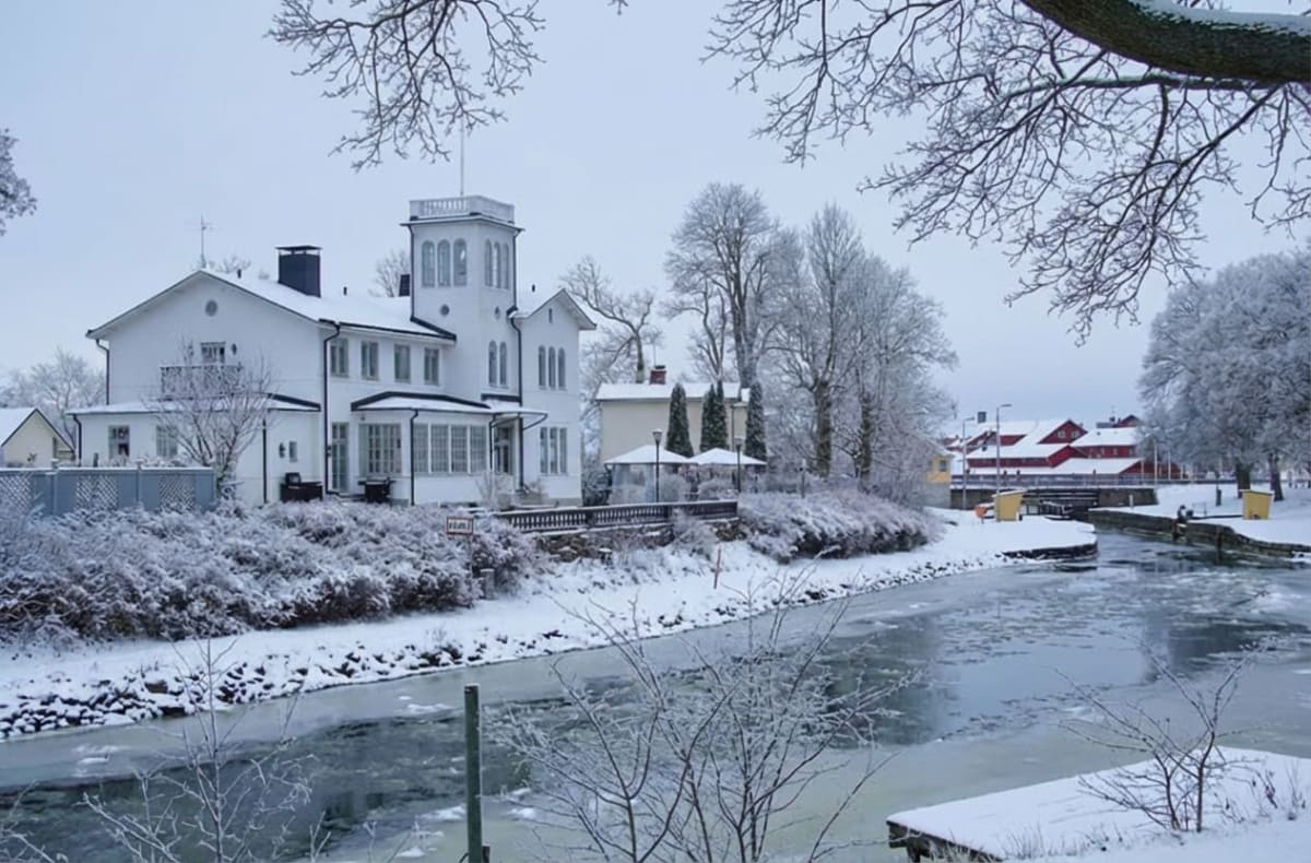 En vinterbild som visar ett delvis fruset vattendrag intill bebyggelse. I förgrunden syns ytan av vattnet med isflak och snötäckta grenar. På vänster sida av vattendraget står en stor, vit byggnad med ett torn och flera fönster, omgiven av snötäckta