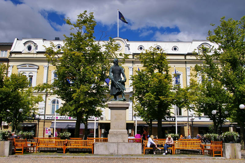 Bilden visar ett torg eller en öppen plats med en bronsstaty föreställande en man på en stenpåt. Statyn är centralt placerad i förgrunden och omges av gröna träd. Bakom statyn och träden skymtar en ljusgul byggnad med vita lister och ett vitt mansard