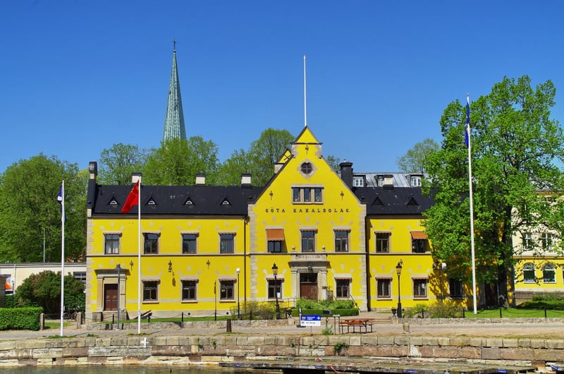 En stor, ljust gul byggnad med tak i mörk färg står i mitten av bilden under en klarblå himmel. På fasaden av byggnaden syns texten "GÖTA KANALBOLAG" under takfoten på den mittersta sektionen. Byggnaden har många fönster med mörka ramar och det finns