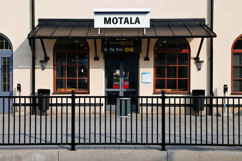 En fasadbild som visar entrén till en byggnad. Ovanför entrédörrarna finns en skylt med texten "MOTALA" och under den en mindre skylt med pilar och symboler som indikerar "Väntsal", "Tåg" och "Buss". Två stora, välvda fönster med spröjs sitter på var