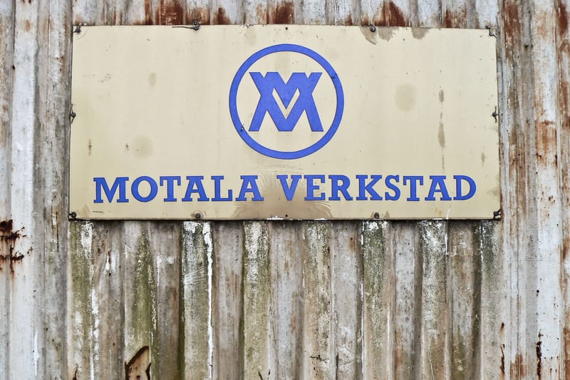 Bilden visar en gulaktig rektangulär skylt med blå text som säger "MOTALA VERKSTAD" och en logotyp ovanför texten. Logotypen består av ett blått, cirkulärt märke med en stiliserad vit bokstavskombination som ser ut som två V:n eller M:n som överlappa