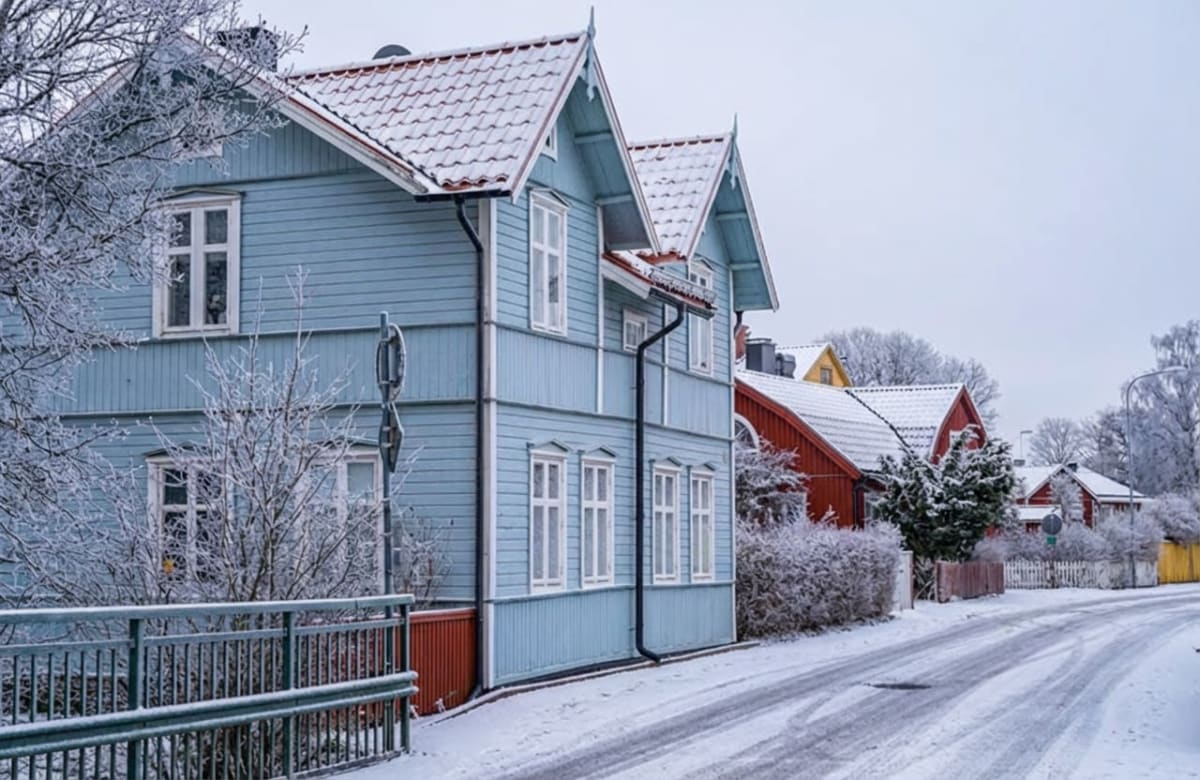 En vinterbild som visar en snötäckt bostadsgata med flera hus. I förgrunden till vänster syns ett ljusblått trähus med vita fönsterkarmar och snö på taket. Huset har en rödbrun grund och en svart stuprör. Framför huset finns ett snötäckt träd och en grå metallräcke. Längs gatan, som är täckt av snö och har spår av fordon, står flera andra hus i olika färger, bland annat röda och gula. Buskar och träd längs vägen är också täckta av snö eller frost. Himmeln är grå och mulen.