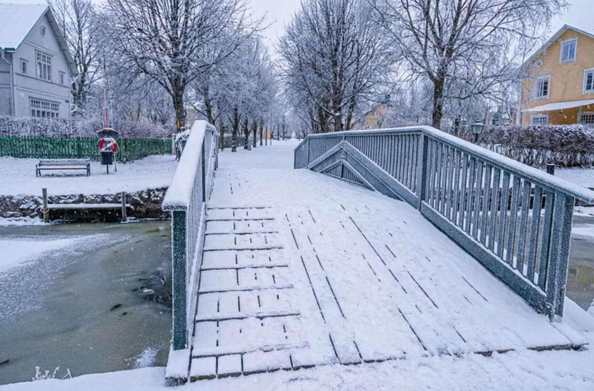 Bilden visar ett snötäckt vinterlandskap med en träbro i förgrunden, som leder över ett delvis isbelagt vattendrag. På brons gångyta och räcken ligger det snö. Till vänster om bron syns en snötäckt gräsyta med en bänk och en röd livboj. I bakgrunden till vänster syns ett vitt hus med mörkgrått tak och vita fönster. Längre bort, längs en gångväg, står flera snöklädda, lövfria träd. Till höger i bilden, bortom bron, syns ett gult hus med vita fönster. Träder är täckta av snö och frost.