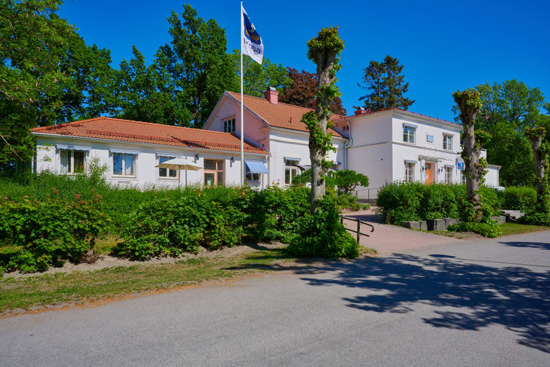 En fasadbild av en större, vit byggnad med tegelrött tak under en klarblå himmel. På byggnadens vänstra sida finns en enplansutbyggnad med flera fönster och en vit solparaply. Huvudbyggnaden är också vit med röda takpannor och har flera fönster med l