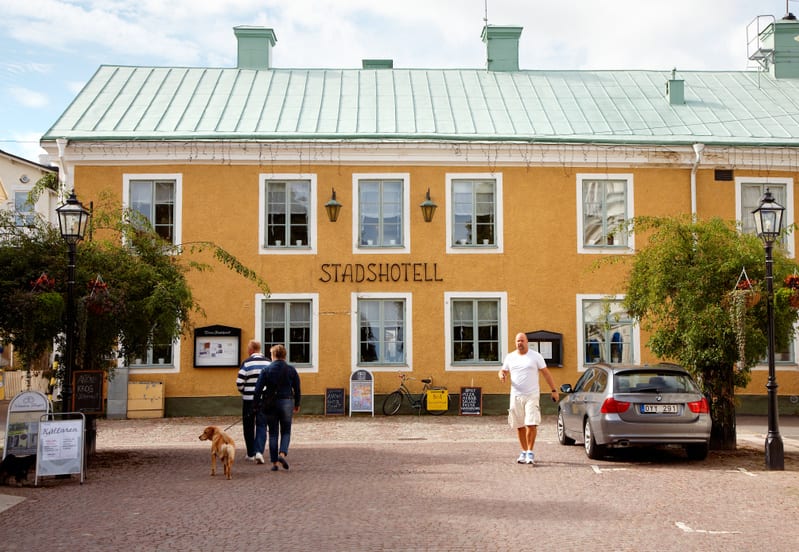 Bilden visar fasaden på en gul byggnad med texten "STADSHOTEL" i mitten. Byggnaden har ett grönt plåttak och flera fönster med vita karmar. Framför byggnaden finns ett kullerstenstorg. En grå kombibil står parkerad till höger i bilden. Till vänster o