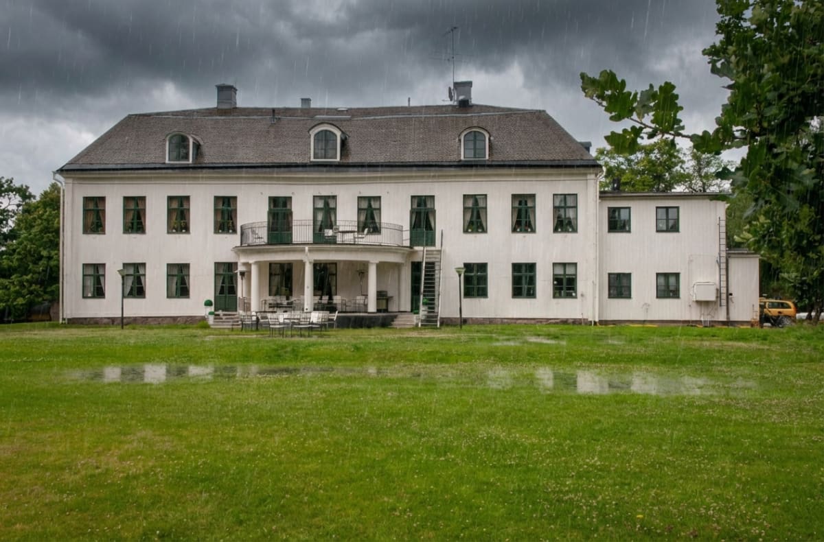 En stor vit byggnad med mörka tak, många fönster och en balkong syns i regnväder. Träd och gräsmattor med vattenpölar omger byggnaden under en gråmulen himmel. En fyrhjuling syns i bildens högra kant.