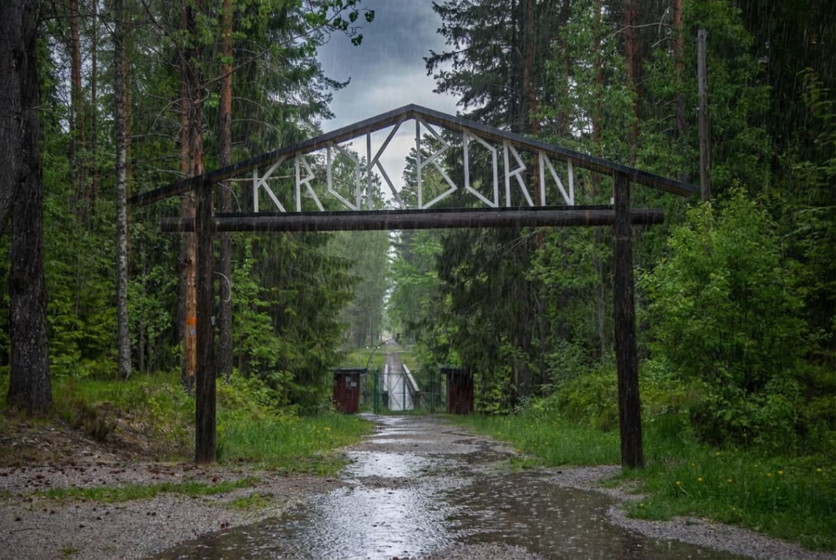 En väg skymtar under en portal med texten "KRÅKBORN" i regnväder. Regndroppar är synliga i luften och på ytor. Vägen sträcker sig mot en öppen grind och en bro, omgiven av träd och vegetation. Himlen är grå och molnig.