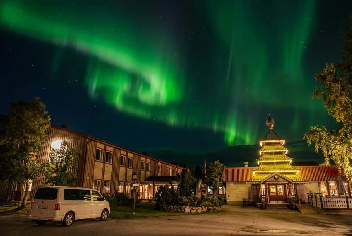 En bild tagen nattetid som visar ett norrsken i gröna nyanser över byggnader. En ljus skåpbil är parkerad framför en av byggnaderna, och träden har gröna löv.