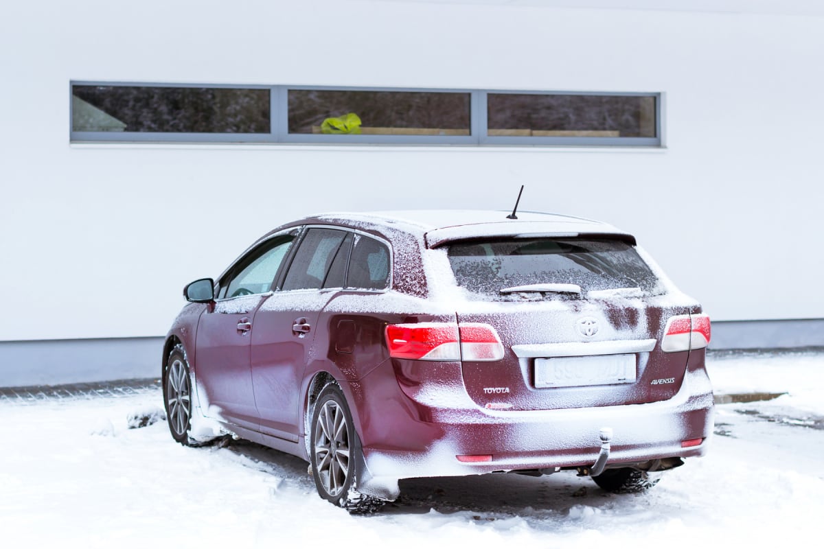 En röd kombibil står parkerad i snö framför en vit byggnad med avlånga fönster. Bilen är delvis täckt av snö, särskilt på taket och bakluckan. I ett av fönstren syns ett gult föremål.