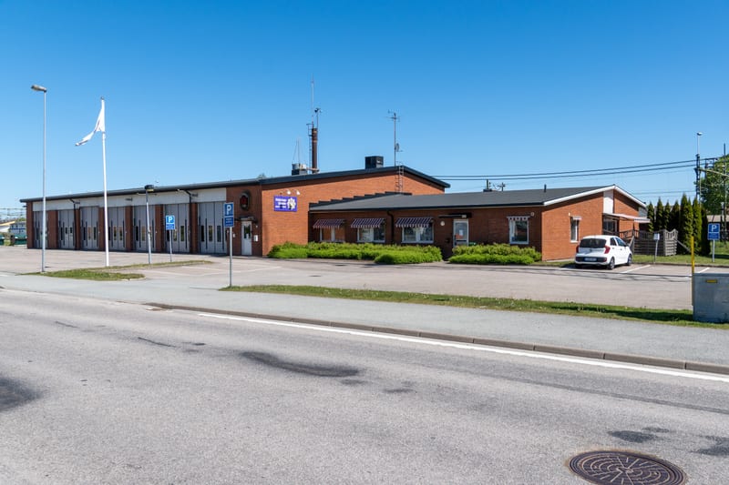 En röd tegelbyggnad, med flera garageportar på vänster sida och fönster med markiser på den högra, står under en klarblå himmel. Framför byggnaden finns en asfalterad yta med parkeringsskyltar och gräsytor. En väg och en brunnslock syns i förgrunden.