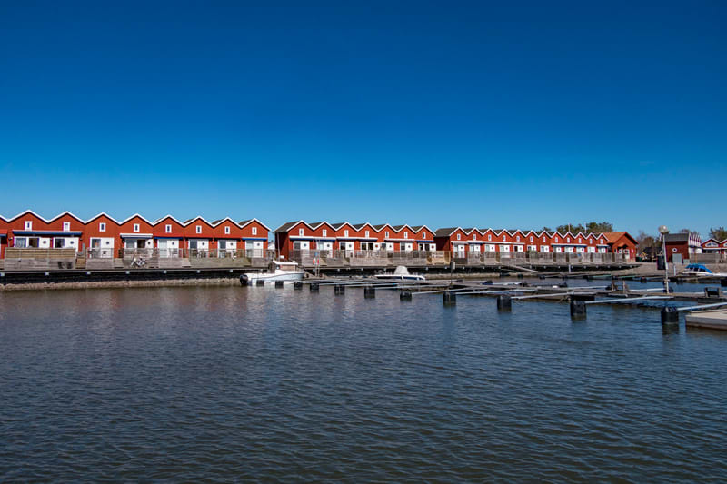 En bild som visar en rad med röda hus med vita detaljer och svarta tak. Husen ligger precis vid en brygga där flera båtplatser syns och ett par båtar är förtöjda. Vattenytan är mörk och himlen är blå och klar.