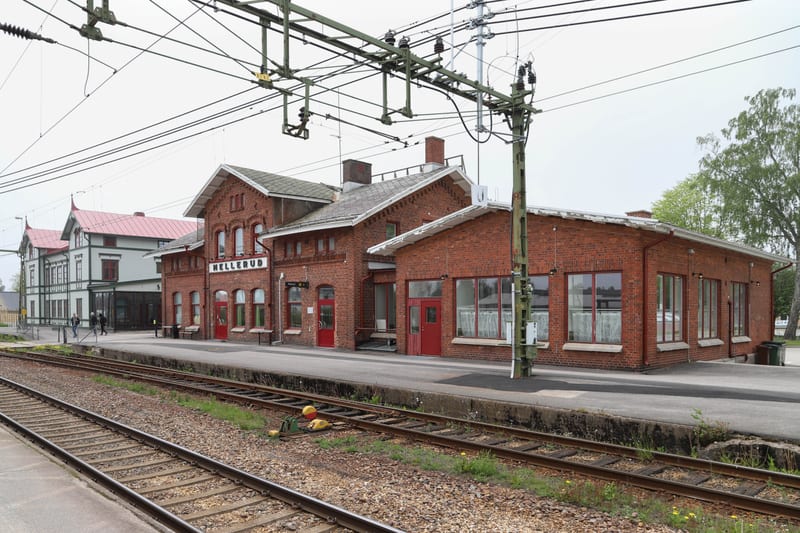 En bild av en tågstation med ett rött tegelhus och texten "MELLERUD" på fasaden, sett från spårsidan. I förgrunden syns tågspår med ett signalsystem. Byggnaden till höger har stora fönster och röda dörrar, medan ett vitt hus med rött tak ses i bakgru