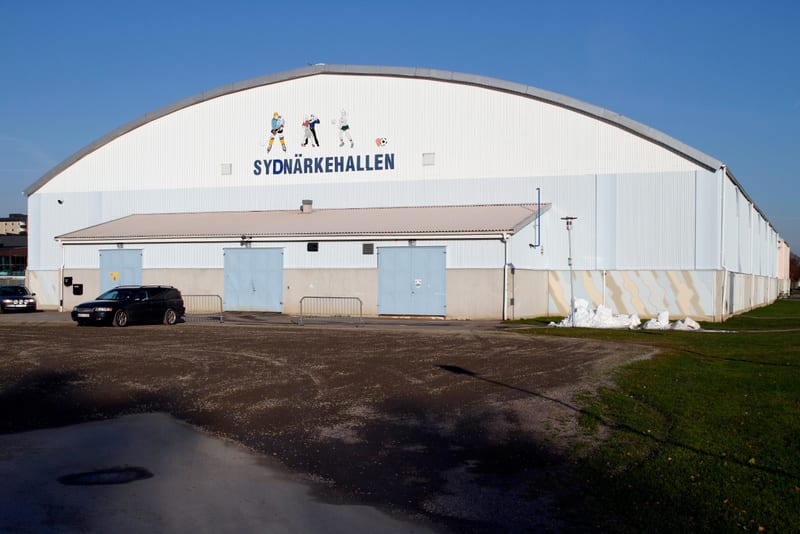 En stor, vit hallbyggnad med krökt tak och texten "SYDNÄRKEHALLEN" samt idrottande figurer på fasaden. Framför hallen finns en grusplan med parkerade bilar och en mindre gräsyta med snöhögar. Blå himmel ovanför.