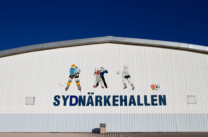 En stor, vit byggnad under en klarblå himmel. På byggnadens fasad syns illustrationer av en ishockeyspelare, två dansande personer och en volleybollspelare. Nedanför illustrationerna står texten "SYDNÄRKEHALLEN" i blåa bokstäver.