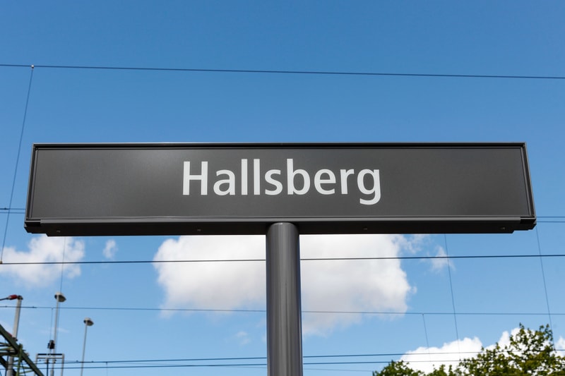 En yttre översiktsbild av en skylt med texten "Hallsberg" i vit färg på en grå bakgrund. Skylten är monterad på en stolpe. I bakgrunden syns blå himmel med några vita moln, samt ledningar och delar av en struktur som kan vara en kontaktledning eller