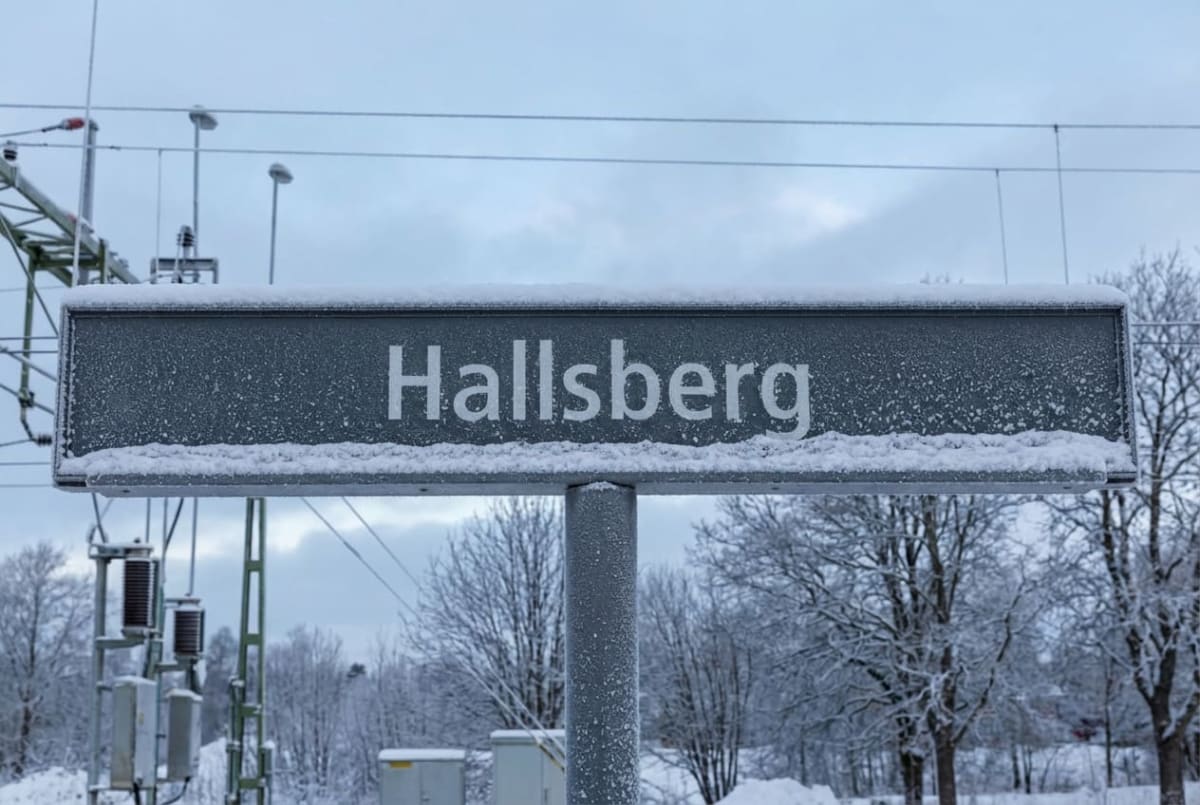 En svart metallskylt med vit text "Hallsberg" är placerad i bildens centrala del. Skylten har snö på sig och står mot en bakgrund av snötäckta träd och stolpar för luftledningar. Himlen är ljus och molnig.