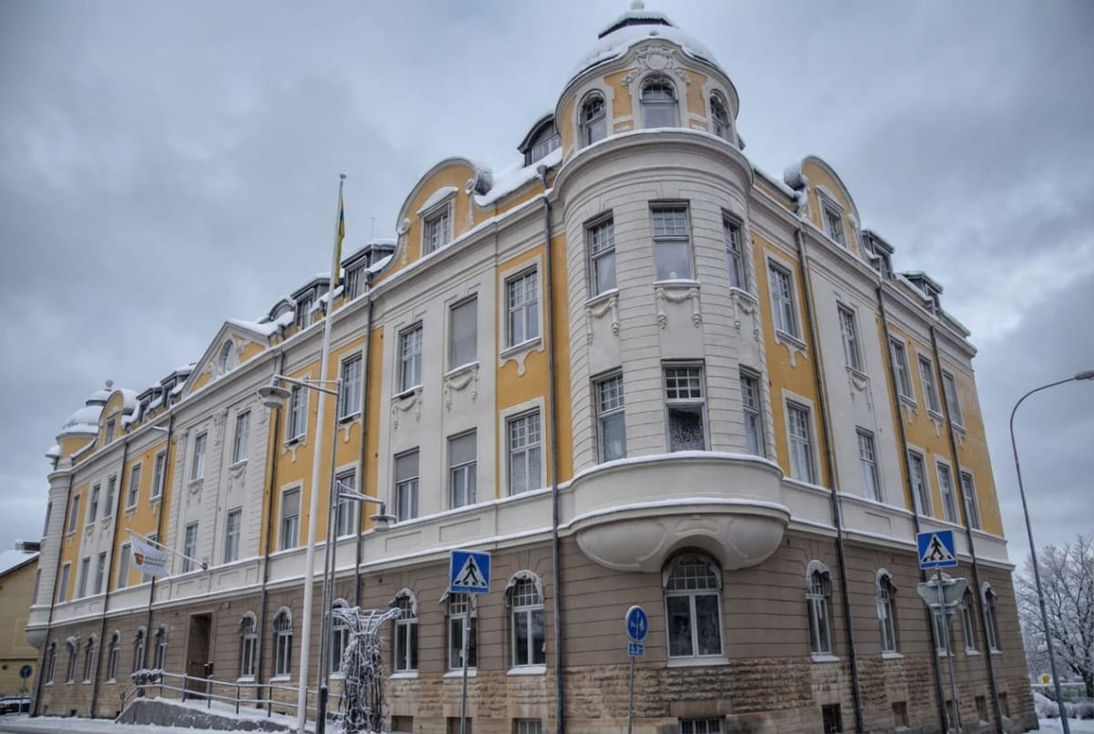 En stor, äldre byggnad med gul och vit puts samt en stensockel. Taket är klätt med snö och har flera fönster längs takfoten. Det finns en flaggstång med en flagga, trafikskyltar, en gatlyktstolpe och en buske nertill, allt täckt med snö.