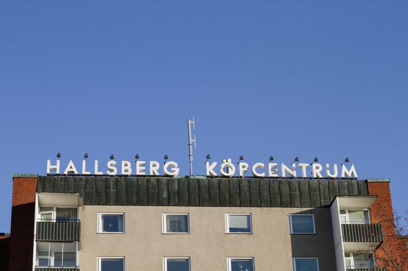 En bild som visar en del av en byggnad med en skylt på taket där det står "HALLSBERG KÖPCENTRUM". Skylten är stor och vit mot en klarblå himmel. Ovanför skylten finns en antenn.