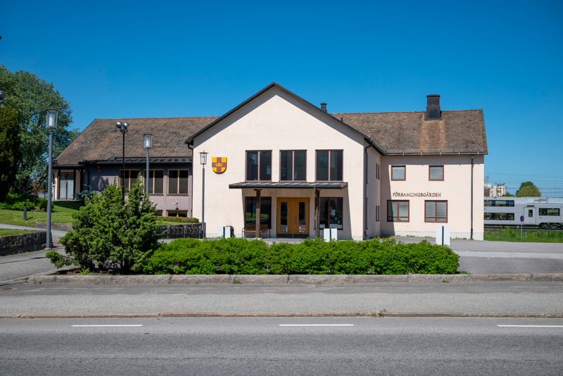 En ljusgrå byggnad med texten "FÖRSAMLINGSGÅRDEN" på fasaden, omgiven av grönskande buskar och träd under en klarblå himmel. Till höger bakom byggnaden syns ett vitt tåg på ett järnvägsspår. I förgrunden syns en asfalterad väg.