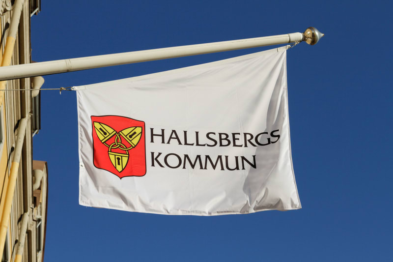 En vit flagga med Hallsbergs kommuns logotyp och texten "HALLSBERGS KOMMUN" hänger från en vit flaggstång mot en klarblå himmel. Logotypen är ett rött fält med tre gula nyckelhål sammanflätade. En del av en gul byggnad syns till vänster.