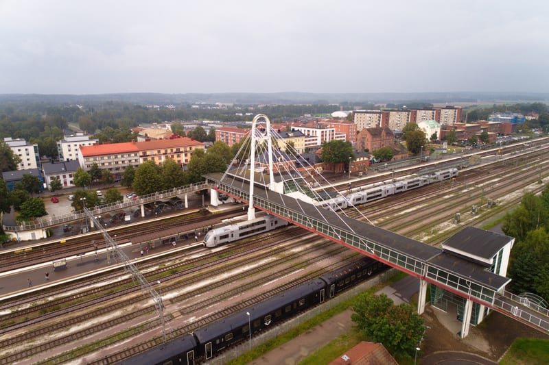 En översiktsbild av ett tågstationområde med flera spår, tåg och en gångbro. I bakgrunden syns stadsbebyggelse med hus och träd, och himlen är molnig. Människor syns på en perrong och en svart byggnad finns i förgrunden.
