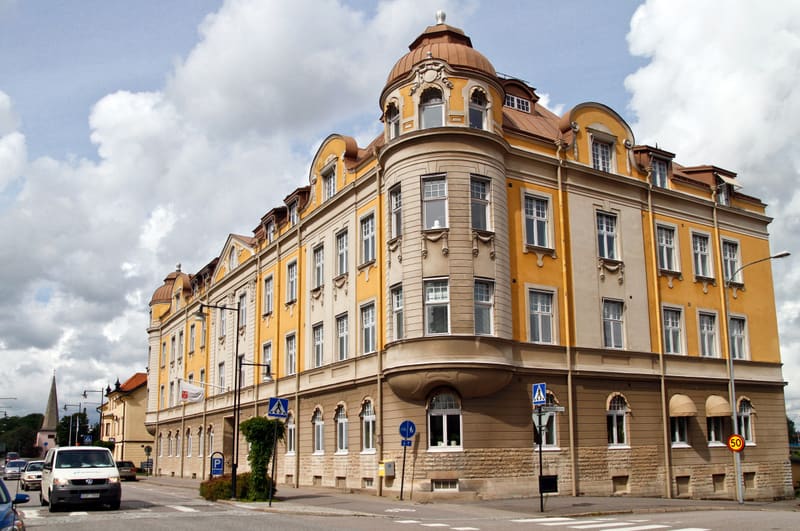 En stor, flerfärgad byggnad av äldre stil dominerar bilden. Byggnaden har detaljerat puts och en kupol på ett hörn. Flera fordon och trafikskyltar syns på gatan framför byggnaden. Himlen är molnig med inslag av blått.