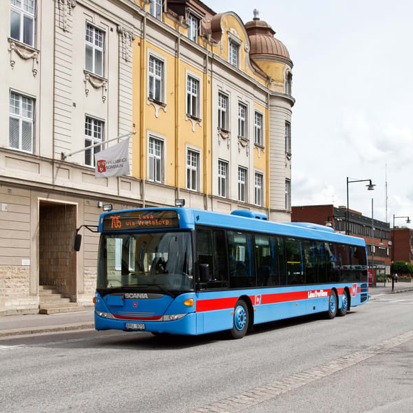 En blå buss med röda ränder kör på en gata framför en gul och vit byggnad. En vit flagga med texten "HALLSBERGS KOMMUN" syns på byggnadens fasad. Bussens destinationsskylt visar "705 Laxå via Vretstorp".