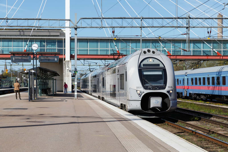 En bild av en tågstation med ett grått passagerartåg på spåret till höger. På plattformen till vänster står två personer. I bakgrunden syns en gångbro med glasväggar och ett blått persontåg. Dagen är solig med en klarblå himmel.