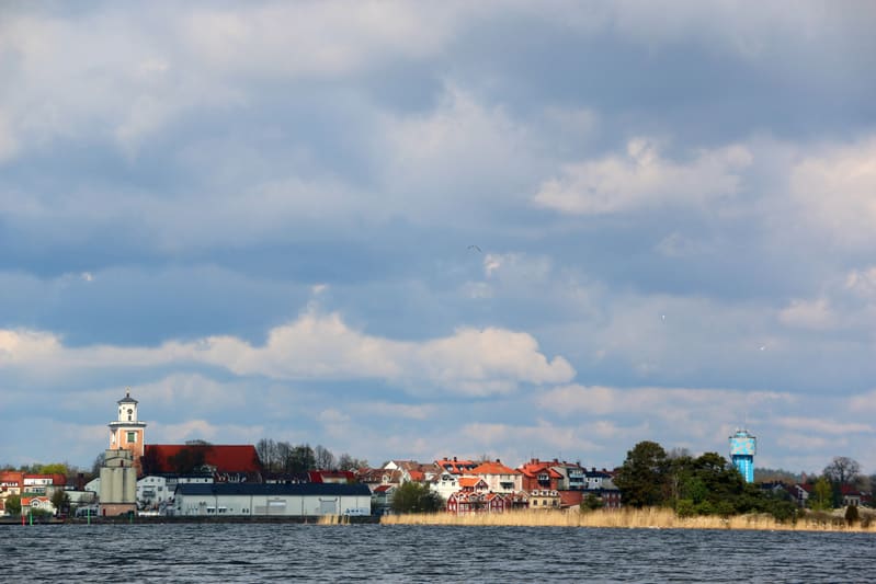 En bild tagen över vatten mot en landremsa med bebyggelse, inkluderande en kyrka med vit kupol och ett blått vattentorn. Bakgrunden är fylld av moln på himlen. I förgrunden syns vattenytan.
