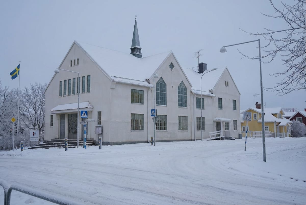En stor, vit byggnad med snötäckt tak och en mindre spira i mitten, omgiven av snötäckta träd och mark. En snötäckt väg slingrar sig framåt, och i bakgrunden syns mindre, färgglada hus. En svensk flagga syns till vänster.