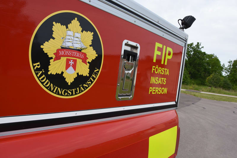 En röd fordonsruta är framträdande i bilden, med texten "FIP FÖRSTA INSATS PERSON" i gula versaler. En logotyp "MÖNSTERÅS RÄDDNINGSTJÄNST" syns också. Bakgrunden består av en asfalterad yta och grönskande träd under en molnig himmel.
