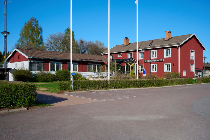 En bild som visar byggnader med röd träfasad och vita knutar under en klarblå himmel. I förgrunden finns en asfalterad parkeringsplats, gröna buskar och en gräsyta. Byggnaderna har bruna tak och på en av dem står det "KOMMUNKONTOR".