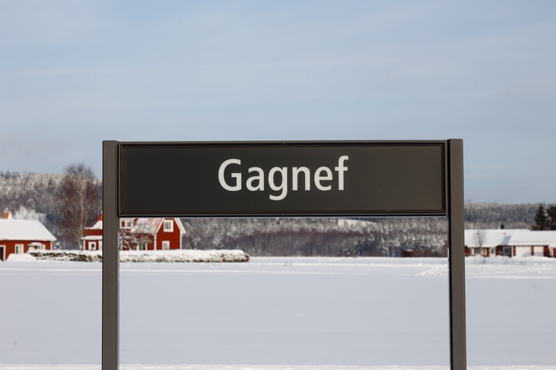 En svart skylt med vit text "Gagnef" står i förgrunden. Bakom skylten syns ett snötäckt landskap med ett fruset vattendrag eller fält, ett antal röda hus och en barrskogsklädd kulle under en klarblå himmel.