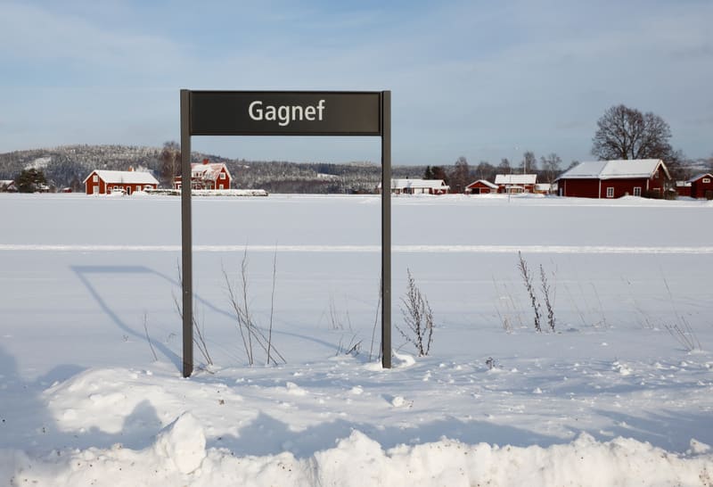 En svart metallskylt med texten "Gagnef" står i ett snötäckt landskap under en klarblå himmel. I bakgrunden syns flera röda byggnader med vita knutar och snötäckta tak, samt snötäckta träd och buskar som sträcker sig mot en kulle. Snö ligger på marke