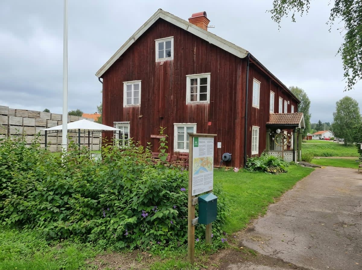 En röd träbyggnad med vita fönsterkanter och tak i förgrunden, omgiven av gräsmattor och buskar. Ett informationsskylt och en grön brevlåda står till vänster om byggnaden, medan en gångväg löper mot höger. I bakgrunden syns träbalkar, en vit paviljon