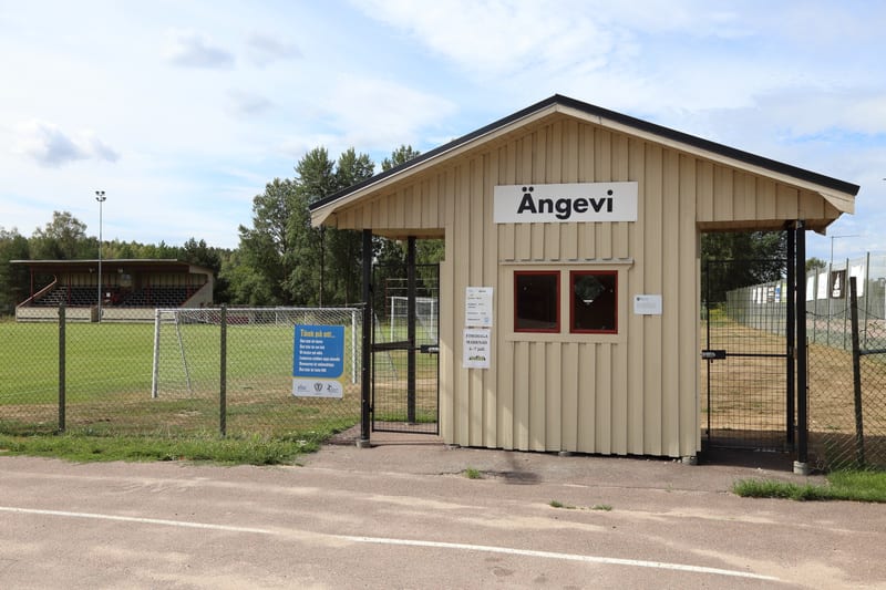 Bilden visar ett fotbollsfält, en läktare och en beige träbyggnad med en skylt märkt "ÄNGEVI". En låst grind och ett staket syns i förgrunden, och i bakgrunden finns träd och en molnig himmel.