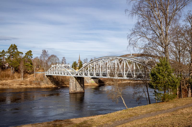 En bro i metall över en flod syns från sidan, med träd och hus på stränderna. Himmelen är ljus med moln. Strandmarken är täckt av brunt torrt gräs. En kyrkspira syns i bakgrunden.
