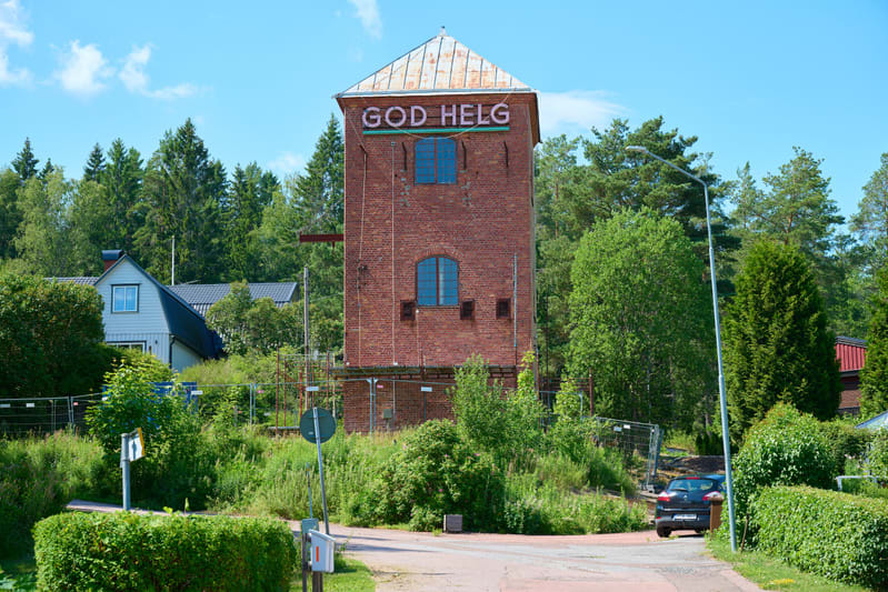 En hög, röd tegelbyggnad med ett metalltak och texten "GOD HELG" på den övre delen, under ett blått underlag. Byggnaden är omgiven av grönskande träd och buskar, med en asfalterad väg och enstaka hus i bakgrunden. En mörk bil syns också i bildens hög