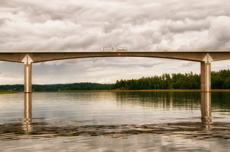 En bro med två långtradare som kör över den. Under bron finns ett avstånd av vatten som reflekterar betongpelaren. Bakgrunden består av en grön skog och en molnig himmel.