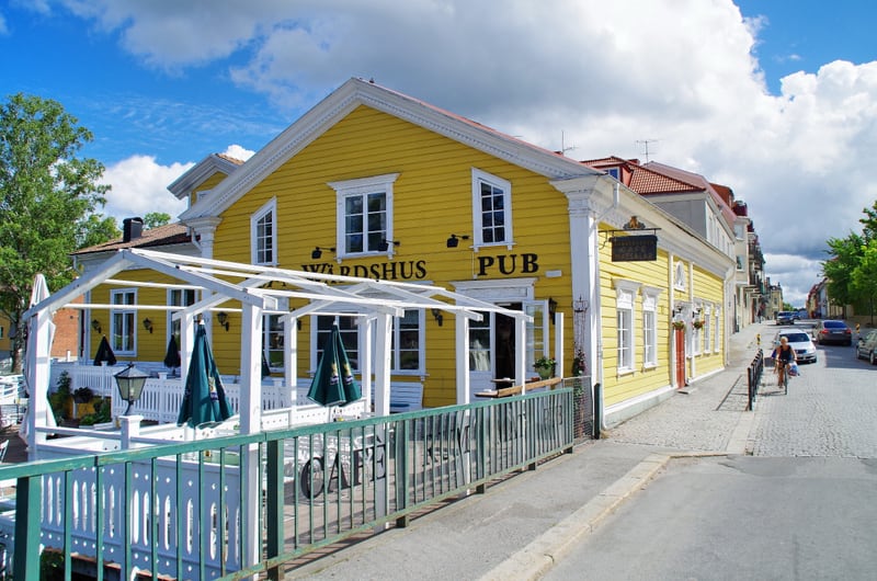 En gul byggnad med texten "WÄRDSHUS PUB" på fasaden står bredvid en gata. Framför byggnaden finns en terrass med vita räcken och gröna parasoller. En person cyklar upp för en gata kantad av parkerade bilar i bakgrunden. Blå himmel med moln ses ovanfö