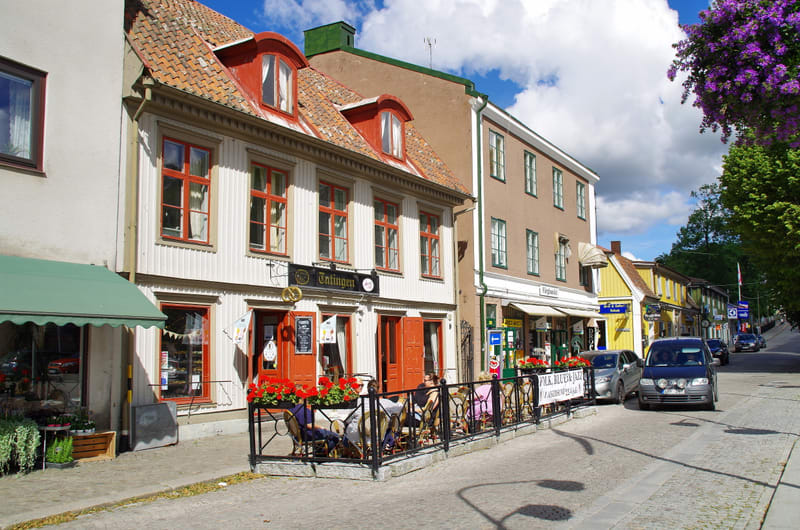 En gata i en stad med en rad hus i olika färger, inklusive en vit byggnad med röda fönsterluckor och en uteservering. Bilparkering syns längs gatan och himlen är blå med vita moln.