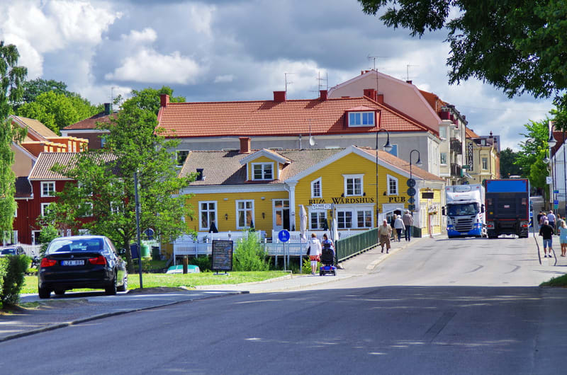 En gata i en stad med byggnader, träd och människor. På vänster sida syns en svart bil parkerad, och längs gatan står byggnader i olika färger, inklusive ett gult hus med skyltar för "Wärdshus" och "Pub". En bro är synlig i förgrunden till höger, med