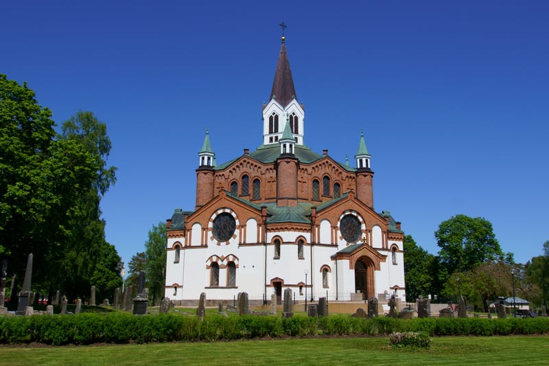 En stor kyrkobyggnad med rött tegel och vita putsade väggar, med flera torn och spetsiga tak, står i centrum av bilden under en klarblå himmel. Framför kyrkan syns en grön gräsmatta och en häck, samt flera gravstenar som antyder en kyrkogård.