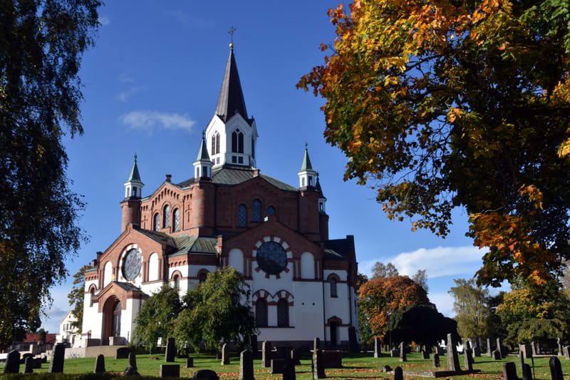 En stor, röd tegelkyrka med vita detaljer och en hög spira, omgiven av en kyrkogård med gravstenar och träd med höstlöv. Himlen är blå med några enstaka moln.