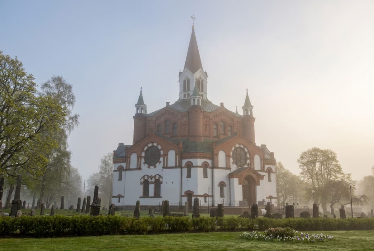 En stor, röd tegelkyrka med vita detaljer och ett högt takspira som reser sig över trädtopparna. Kyrkan är omgiven av en kyrkogård med gravstenar och grön gräsmatta. Dimma ligger över landskapet och förmedlar en lugn, stämningsfull känsla.