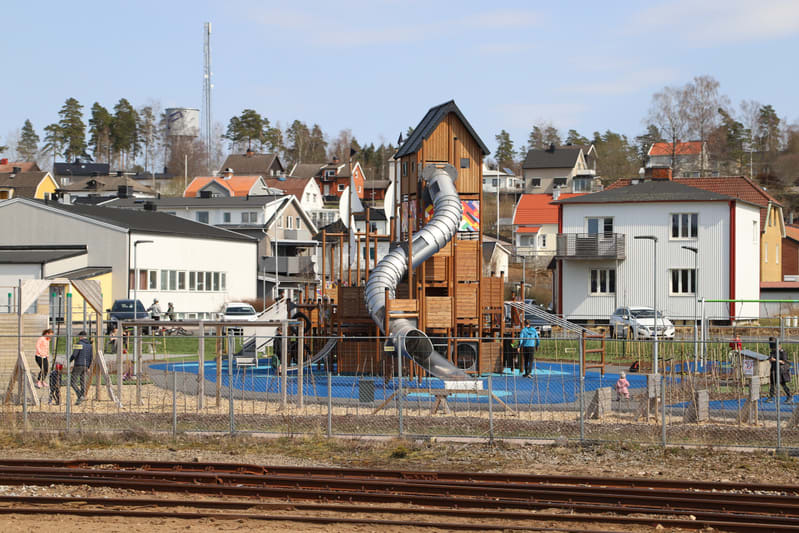 En sandfärgad staket avgränsar framsidan av bilden, med tågspår synliga i dess nedre del. Bakom staketet finns en lekplats med en stor träkonstruktion med en cylinderformad rutschkana, en blå matta och flera vuxna och barn. Bakom lekplatsen syns bost
