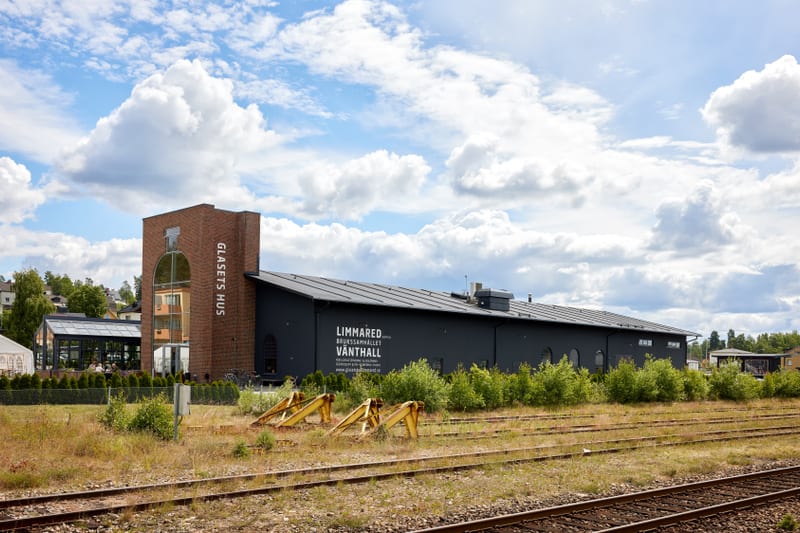 En bild visar två byggnader, en tegelfärgad och en mörkgrå, i ett område med gräs och tågspår. Den tegelfärgade byggnaden har stora fönster och texten "GLASETS HUS". Den mörkgrå byggnaden, som har solpaneler på taket, har en skylt med texten "LIMMARE