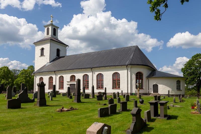 En vit kyrkobyggnad med grått tak och en fyrkantig torn med en guldkors på toppen, omgiven av en grön kyrkogård med gravstenar. Blå himmel med vita moln syns ovanför kyrkan och träd. Rödfärgade fönsterramar.
