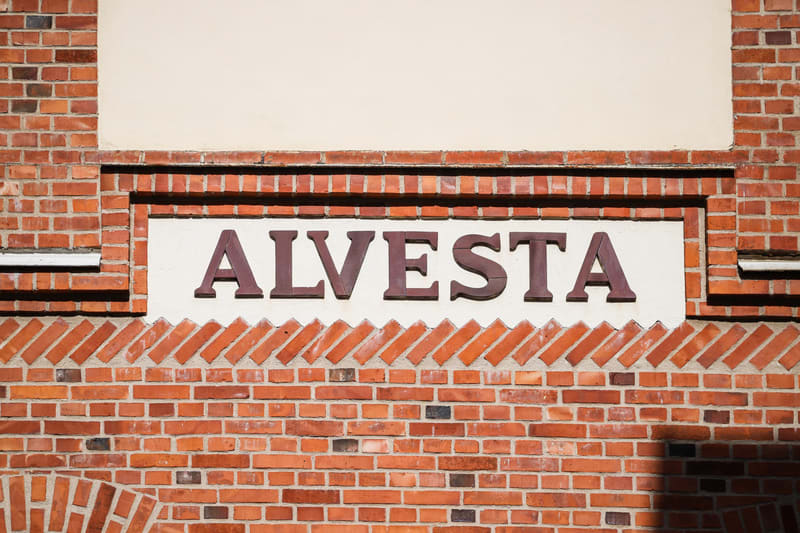 En närbild av en tegelfasad med ordet "ALVESTA" i mörkbruna, upphöjda bokstäver på en ljus skylt. Teglet är rödaktigt med mörkare fogar, och det finns både horisontella och diagonala tegelmönster.